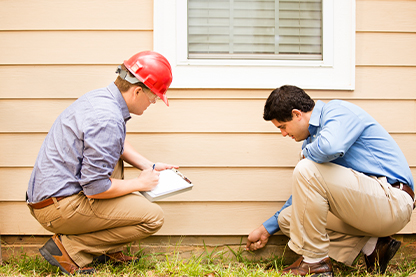 Image: Two men kneeling and pointing to a house foundation. One man has a hard hat and clipboard.